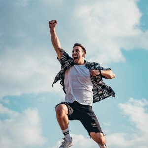 A joyful young man jumps midair with clouds and blue sky in the background, exuding energy and freedom.